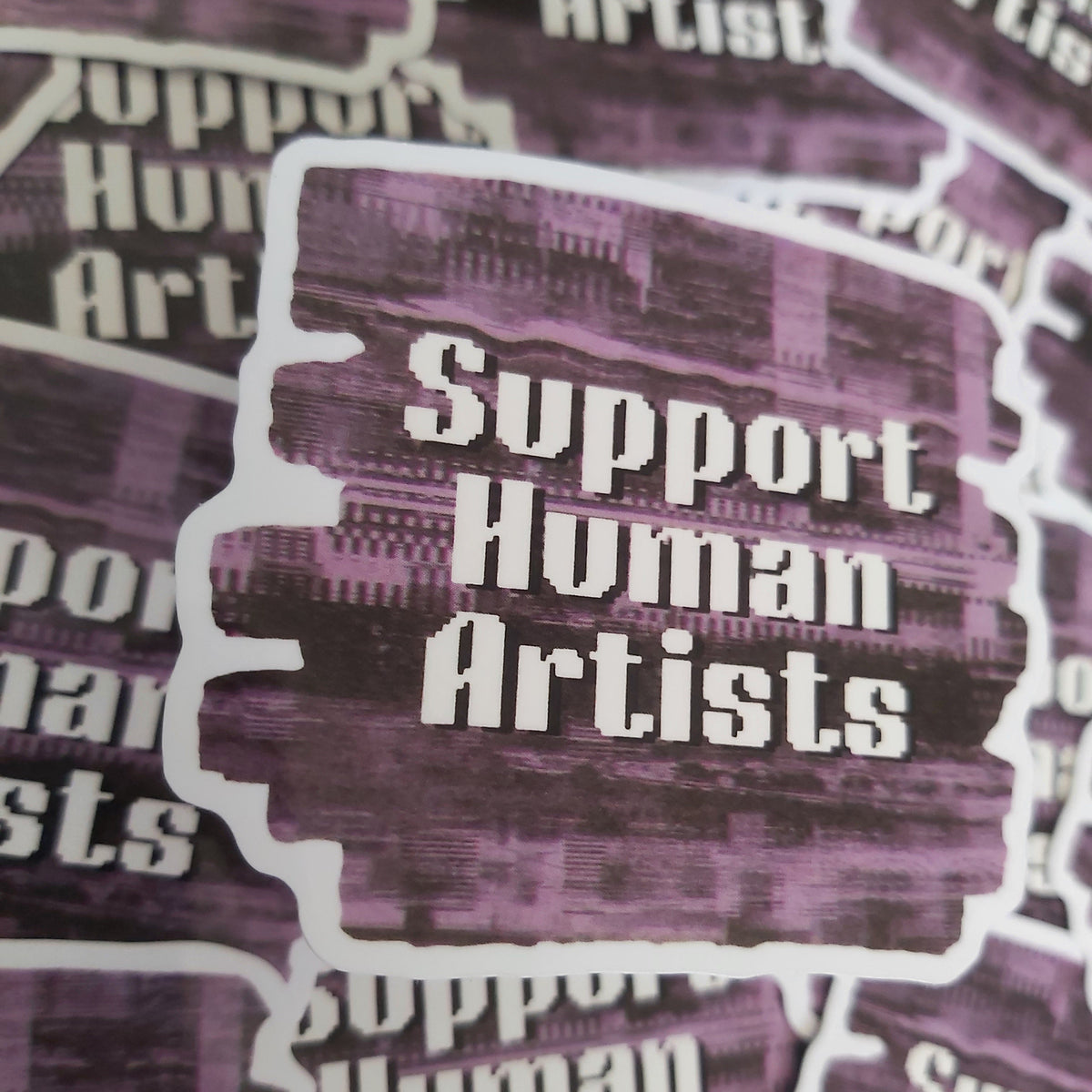 A close-up of a die-cut sticker with the words 'Support Human Artists' in white text over a purple and black textured background, with additional copies of the sticker visible around it.