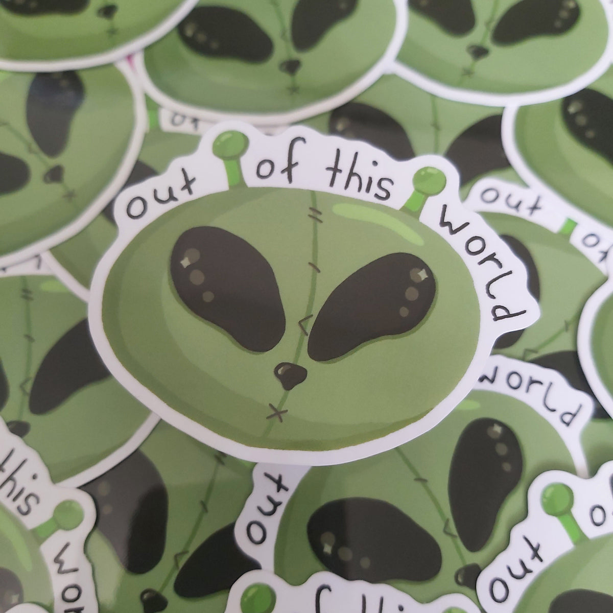 A pile of die-cut stickers featuring a green alien head with black eyes and the phrase 'out of this world' in white text, with one sticker centred in the foreground.