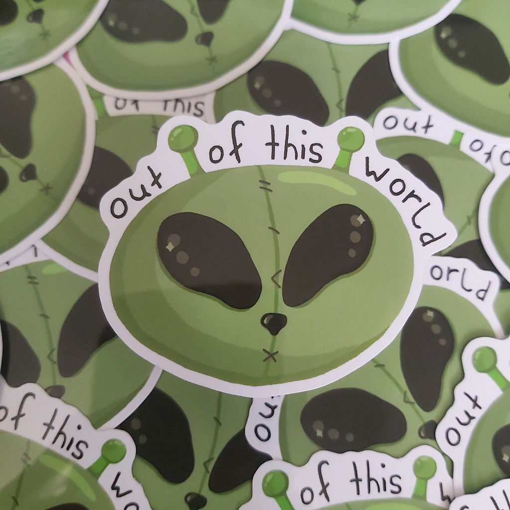 A close-up of a pile of die-cut stickers featuring a green alien head with black eyes and the phrase 'out of this world' in white lettering on a black background.