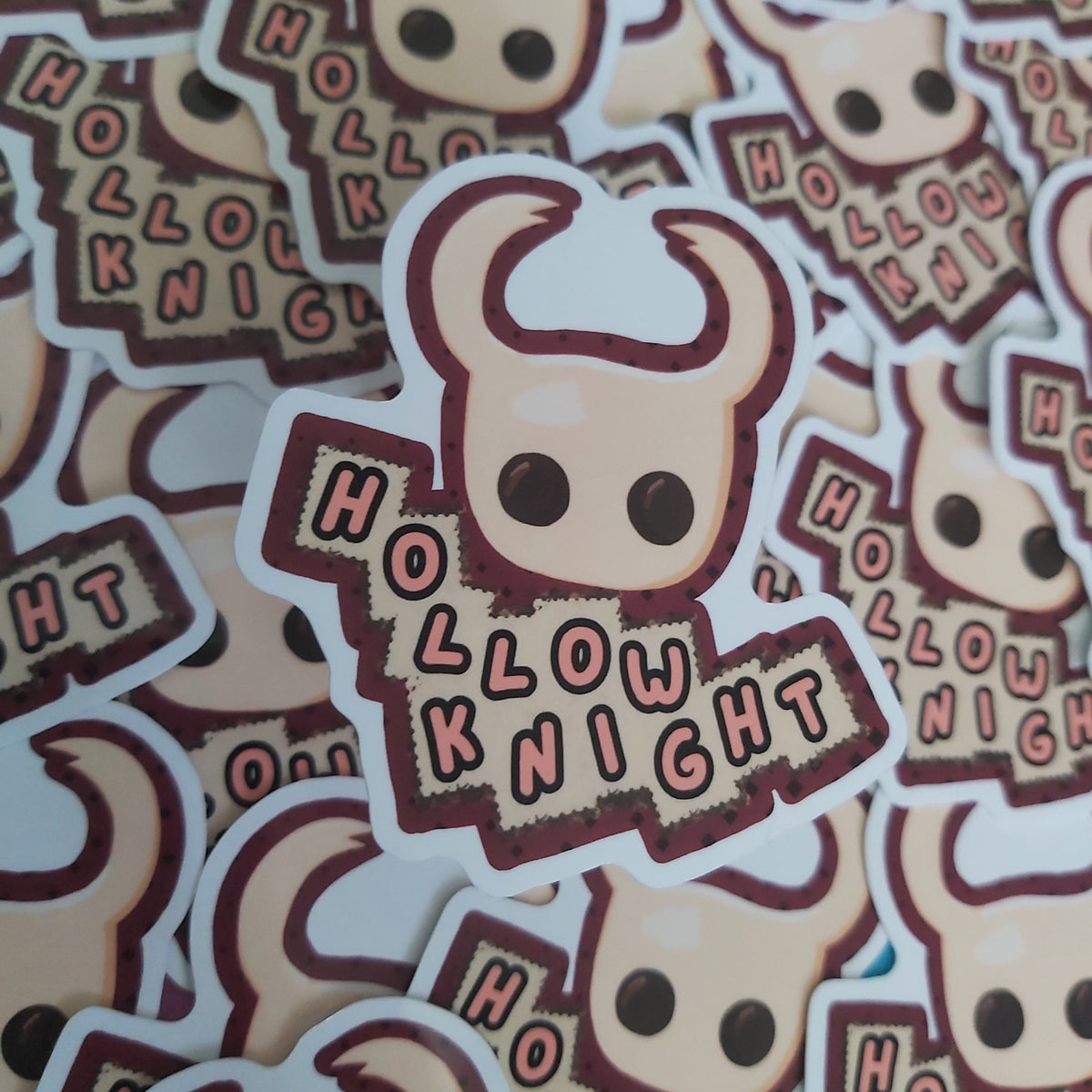 A pile of die-cut stickers featuring a cartoon horned knight head and the words 'HOLLOW KNIGHT' in a stylized font, photographed close up.