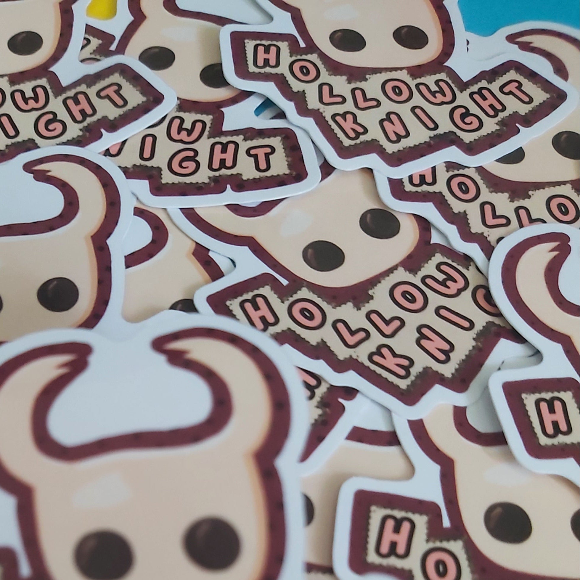 A pile of die-cut stickers featuring a cartoon knight head with horns and the words 'HOLLOW KNIGHT' in pink lettering on a dark brown background.