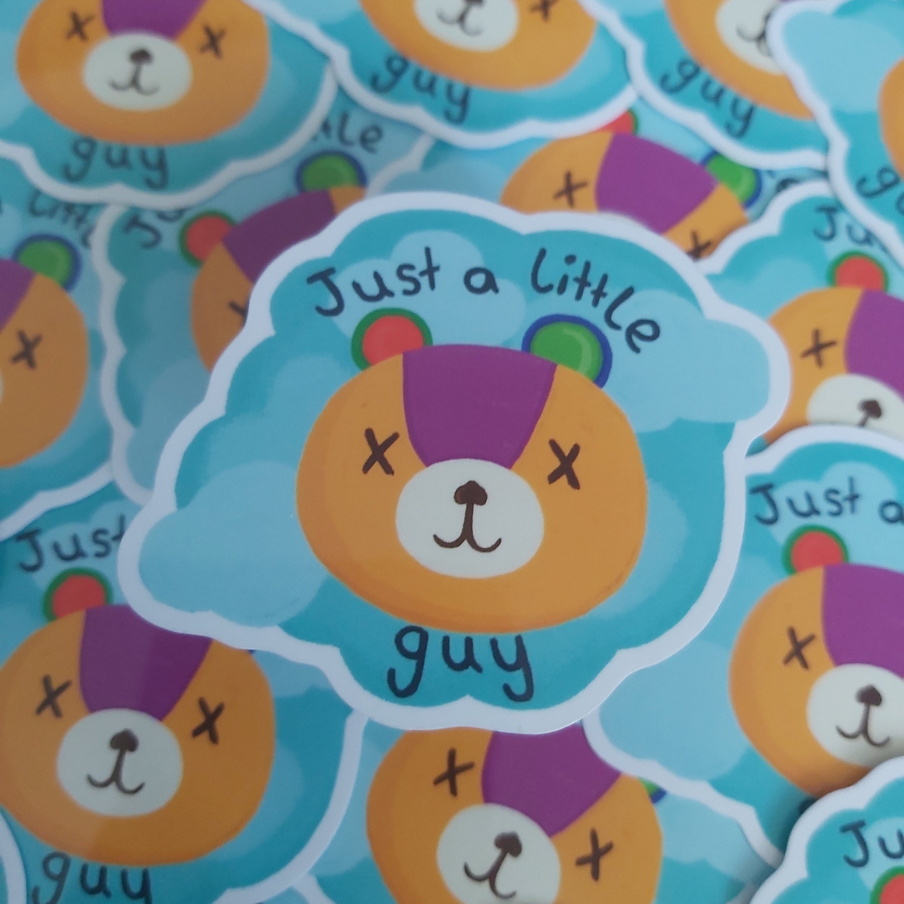 A close-up of a stack of die-cut stickers featuring a cartoon teddy bear face with the text 'Just a little guy'. The sticker has a light blue cloud-like outline, an orange bear face,  and black text.