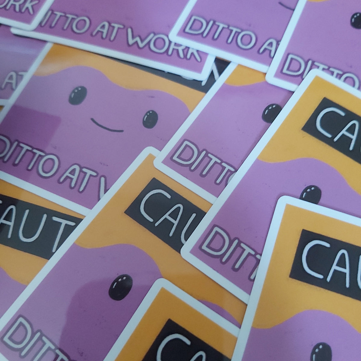 A close-up of multiple cartoon stickers arranged diagonally. The stickers feature a purple character with a simple smiley face on a yellow background, alongside orange and black warning-style labels with white text.