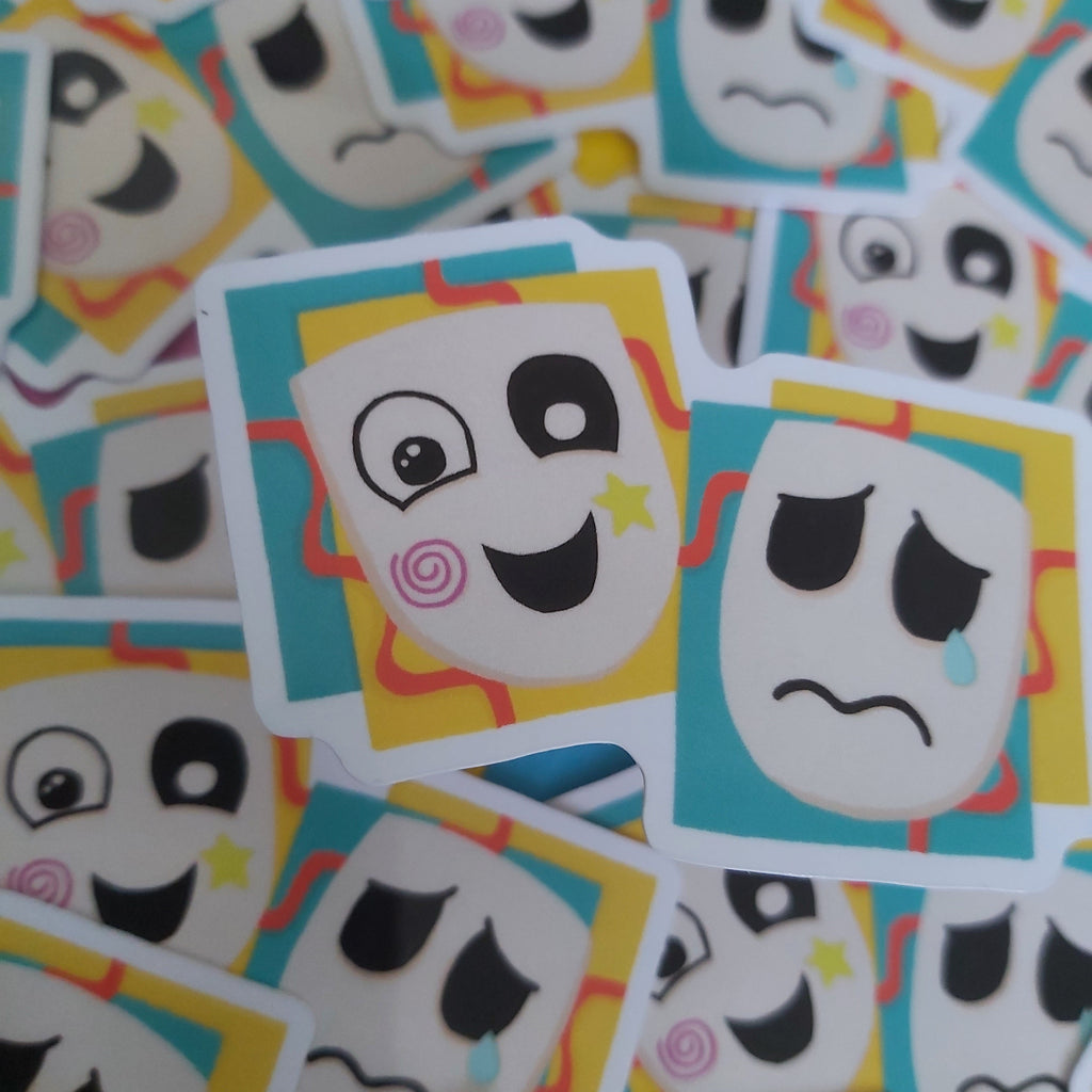 A pile of square stickers featuring cartoon theatre masks. The left side shows a smiling comedy mask with a pink swirl on its cheek, and the right side has a sad tragedy mask with a tear.