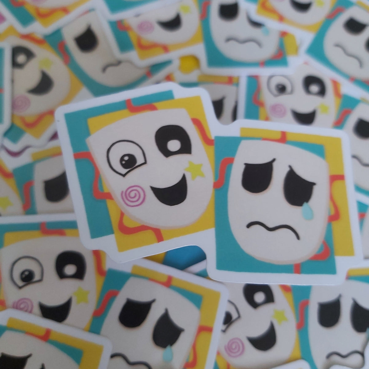 A pile of small square stickers featuring cartoon faces in black, white, yellow, teal, and pink, with several stickers in the foreground in sharper focus.