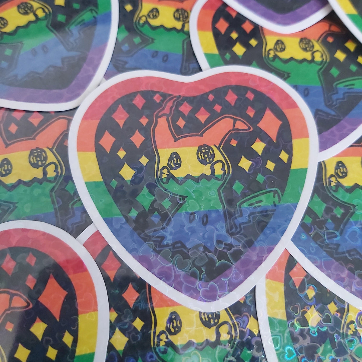 A close-up of heart-shaped stickers with a rainbow background and a black silhouette of a Pokémon character illustration, overlapping on a white background.