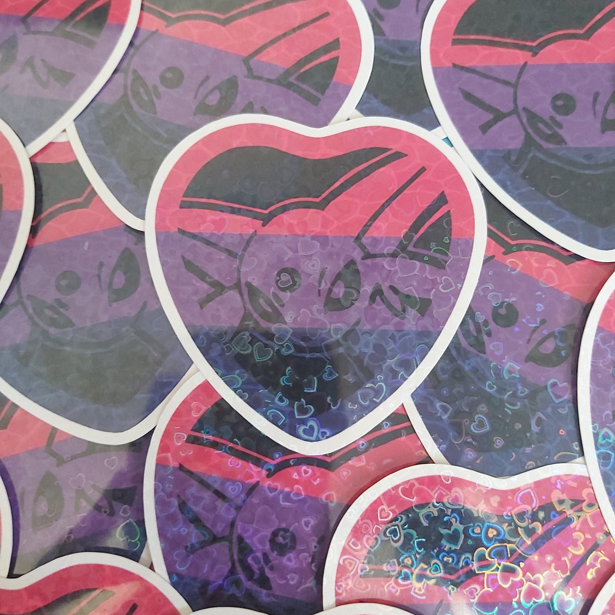 A pile of heart-shaped stickers featuring a stylised Pokémon face layered over horizontal pink, purple, and blue stripes, with holographic glitter accents on some stickers.