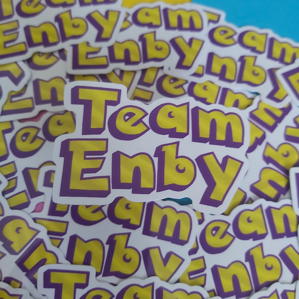 A pile of die-cut stickers featuring the text 'Team Enby' in yellow letters with a purple outline on a white background, photographed close-up against a split yellow and blue backdrop.