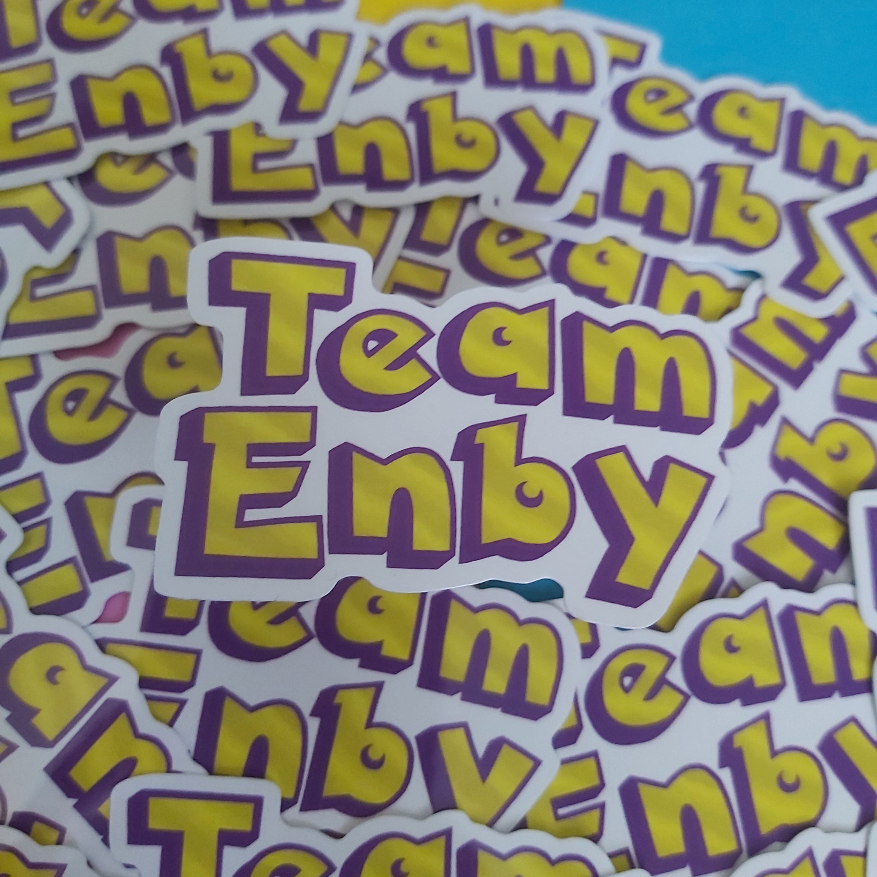 A pile of die-cut stickers featuring the text 'Team Enby' in yellow letters with a purple outline on a white background, photographed close-up against a split yellow and blue backdrop.