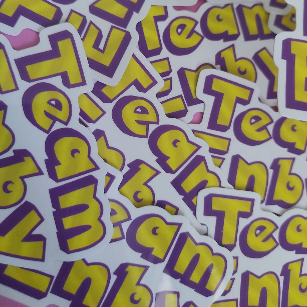 A pile of die-cut stickers featuring the word 'Team ENBY' in bold yellow letters with a purple outline on a white background, scattered across a pink surface.