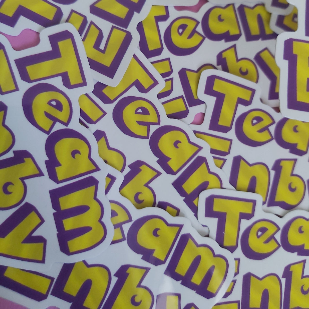 A pile of die-cut stickers featuring the word 'Team ENBY' in bold yellow letters with a purple outline on a white background, scattered across a pink surface.