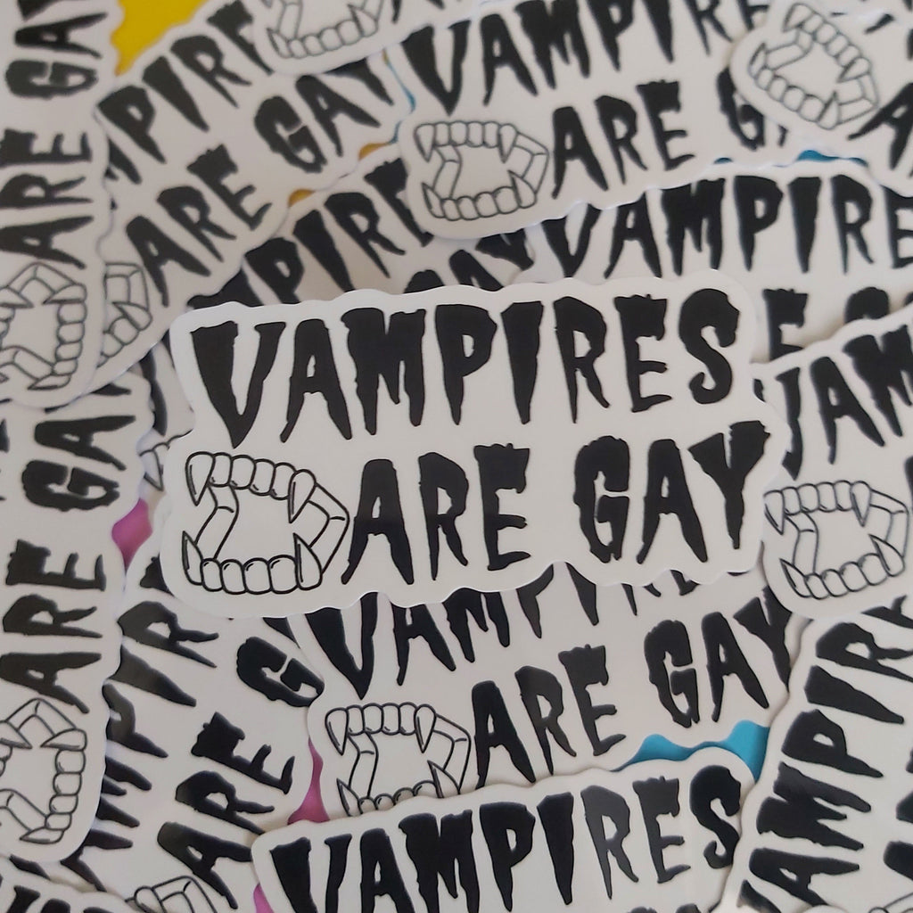 A pile of white die-cut stickers featuring the text 'VAMPIRES ARE GAY' in a black gothic font, accompanied by illustrations of vampire fangs, scattered over a background of matching stickers.