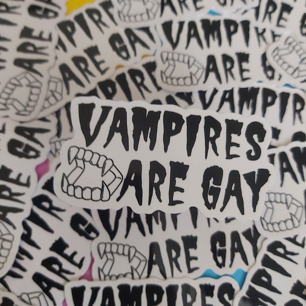 A pile of white stickers with black text reading 'VAMPIRES ARE GAY' and black line drawings of vampire fangs, photographed close up against a bright yellow and blue background.