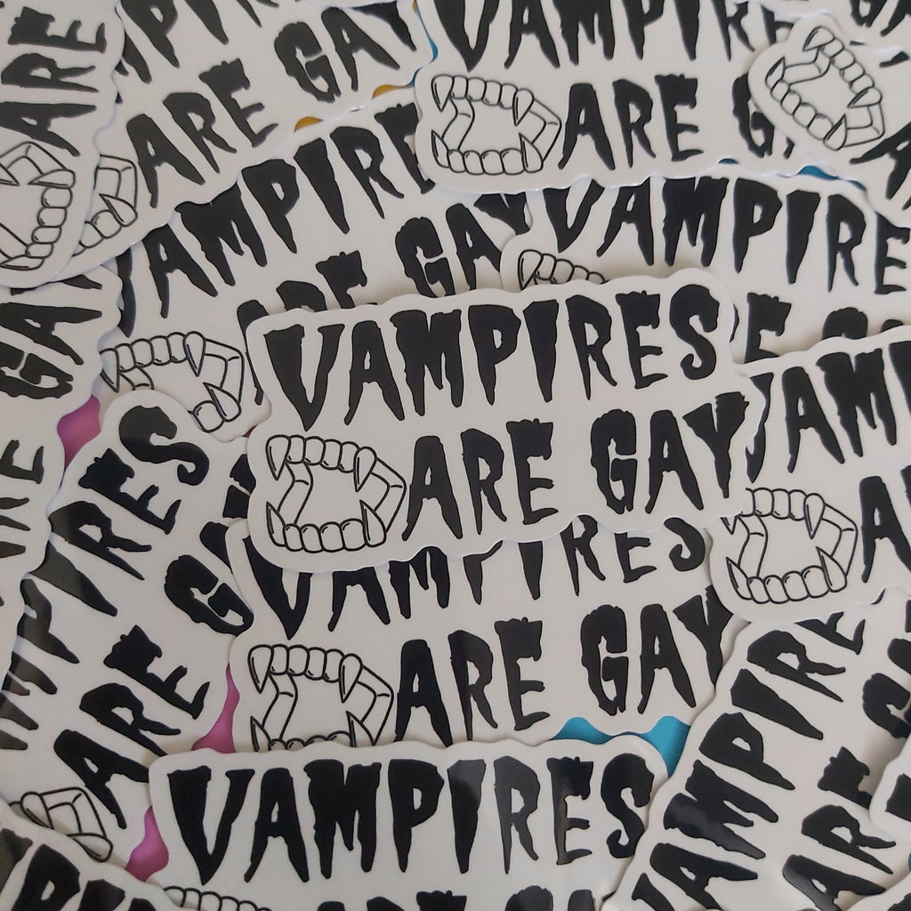 A pile of white die-cut stickers featuring the phrase 'VAMPIRES ARE GAY' in a black gothic font, accompanied by black line drawings of vampire fangs.