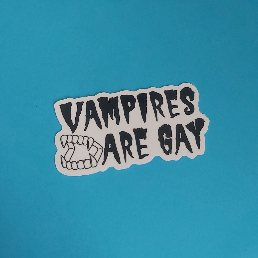 A die-cut vinyl sticker with the text 'VAMPIRES ARE GAY' in a black hand-drawn font next to a black line art illustration of vampire fangs, shown on a solid blue background.