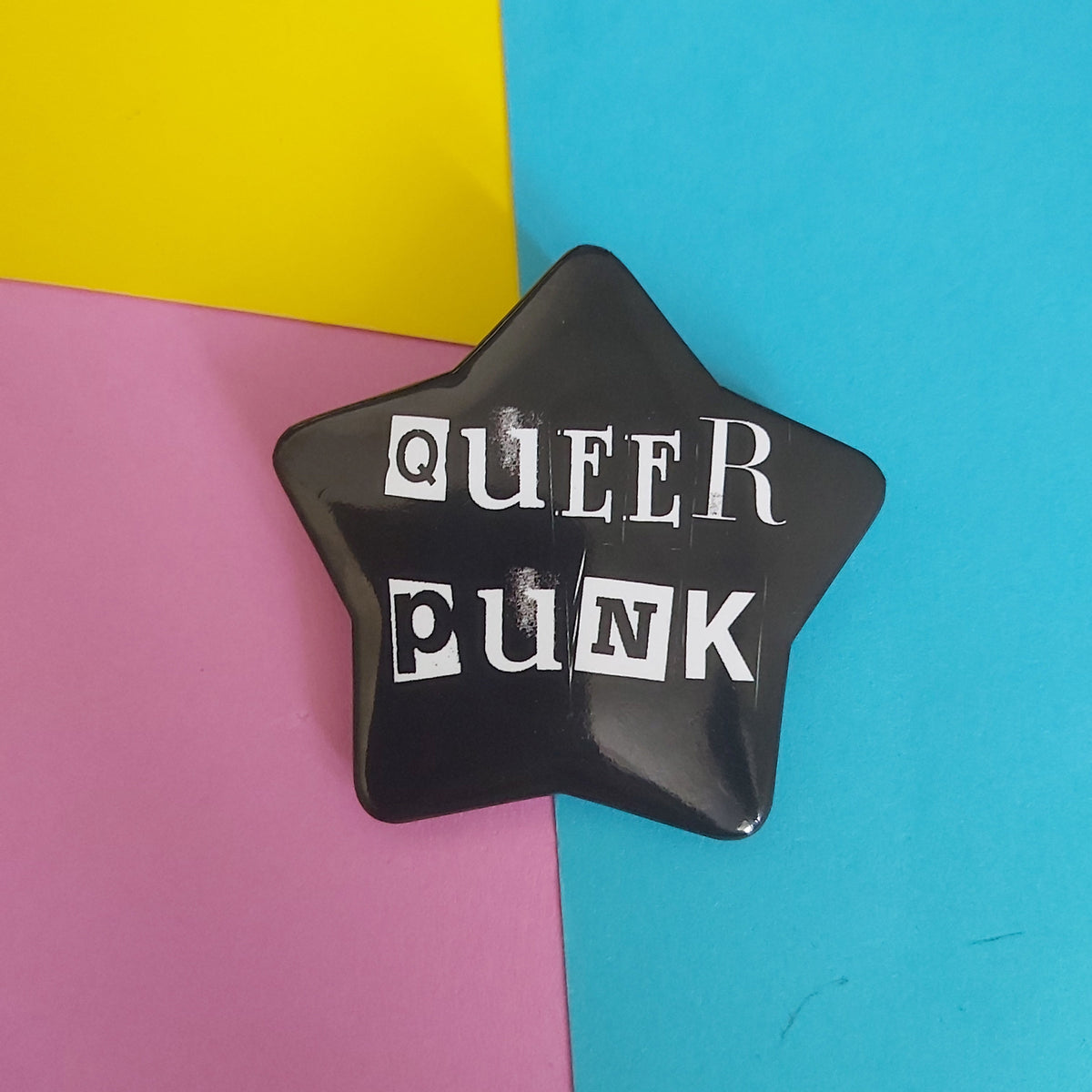 A glossy black star-shaped pin with the words 'QUEER PUNK' in white block letters, displayed on a background of three colored paper sections: yellow, pink, and blue.