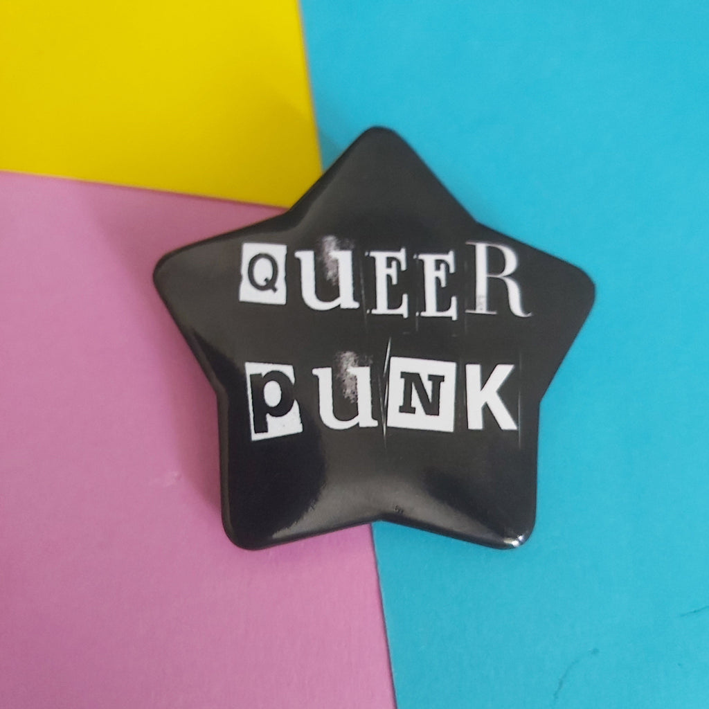 A glossy black star-shaped pin with the words 'QUEER PUNK' in white block letters, displayed on a colourful background of yellow, pink, and blue paper.