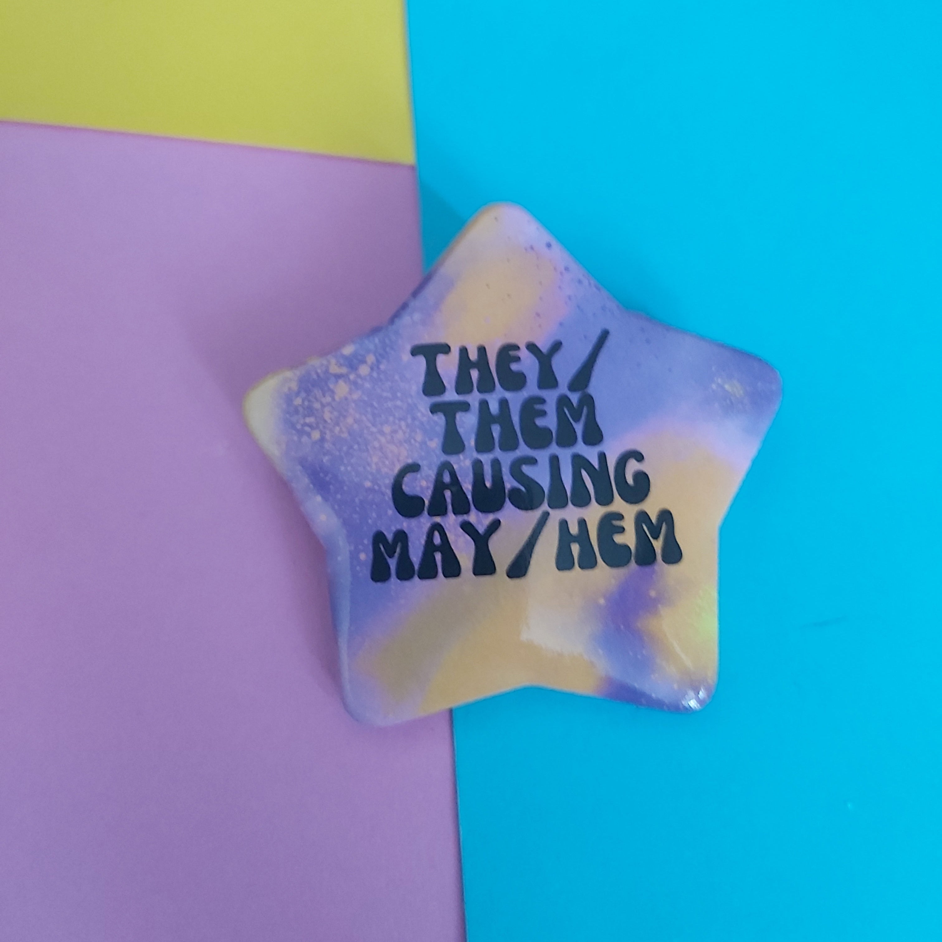 A star-shaped pin with a purple, yellow, galaxy-like marbled pattern, featuring the hand-lettered text 'THEY / THEM CAUSING MAY/HEM' in black. The pin is photographed on a background split diagonally between pastel purple and bright cyan blue.