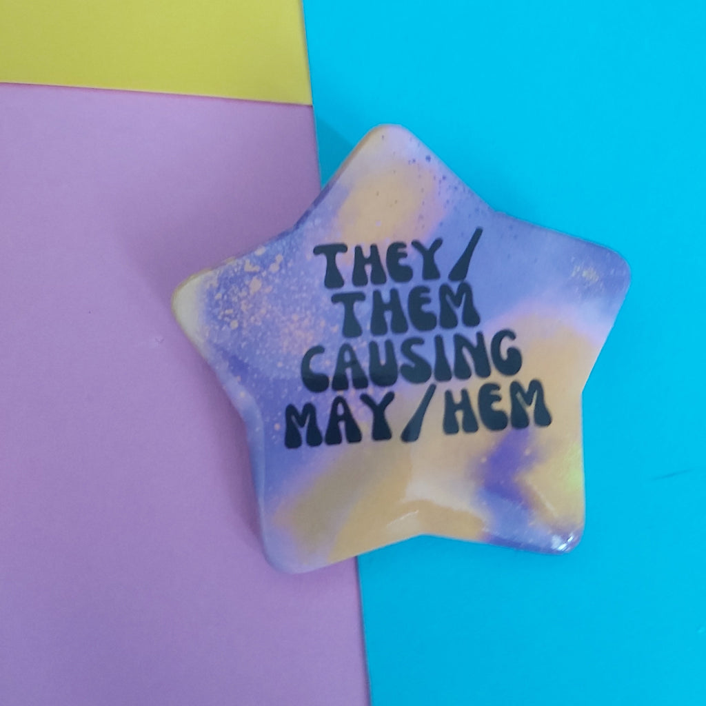A star-shaped pin with a marbled pastel pattern in purple and yellow featuring the text 'THEY/ THEM CAUSING MAY/HEM' in black. The pin is displayed on a background split diagonally between pink and bright cyan blue.
