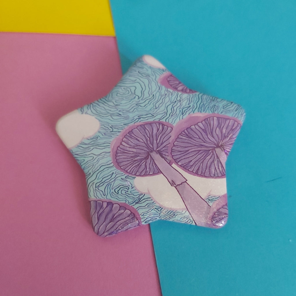 A star-shaped pin badge featuring a purple and light blue mushroom pattern on a white background, displayed on a colourful background of pink, yellow, and blue paper.