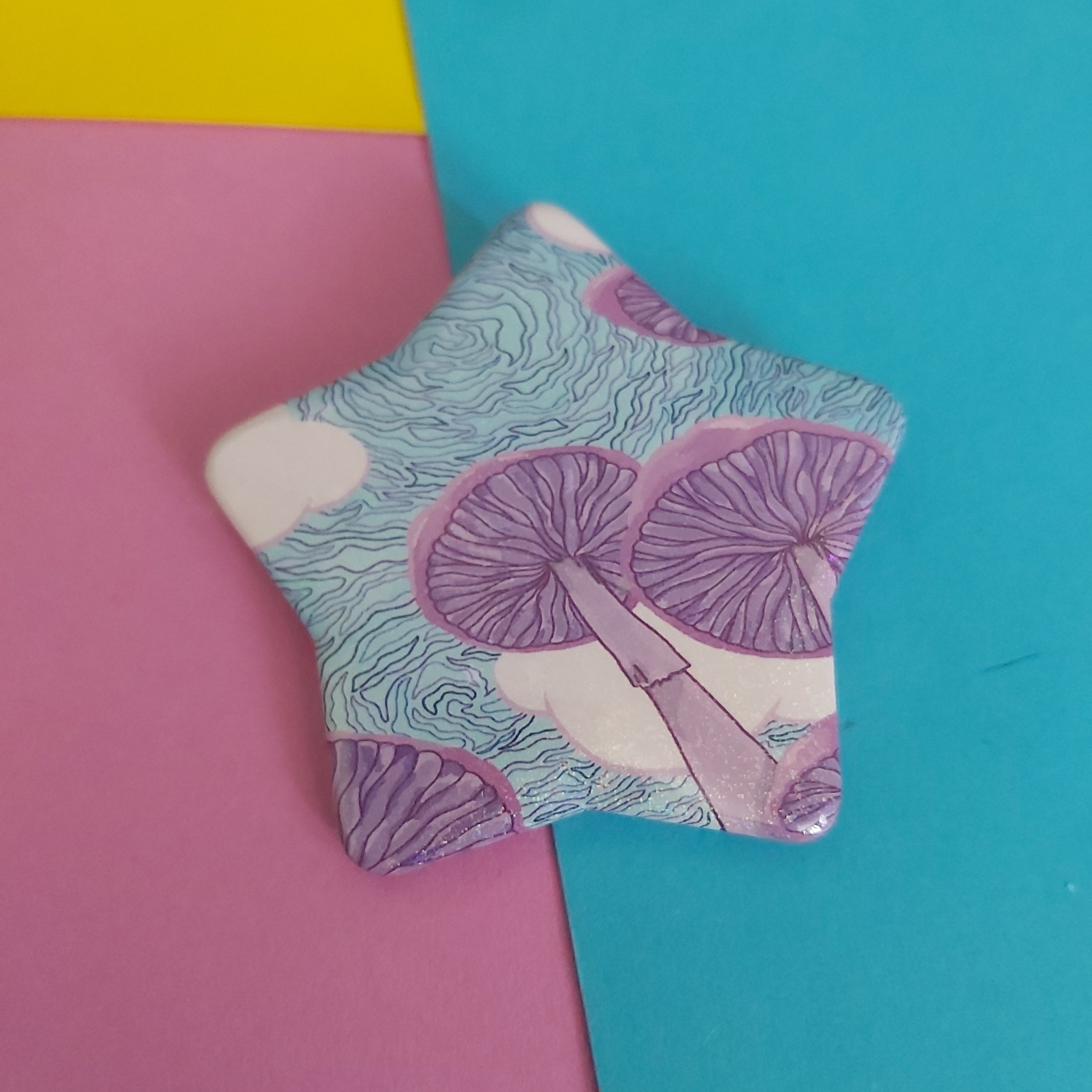 A star-shaped pin badge featuring a purple and light blue mushroom pattern on a white background, displayed on a colourful background of pink, yellow, and blue paper.