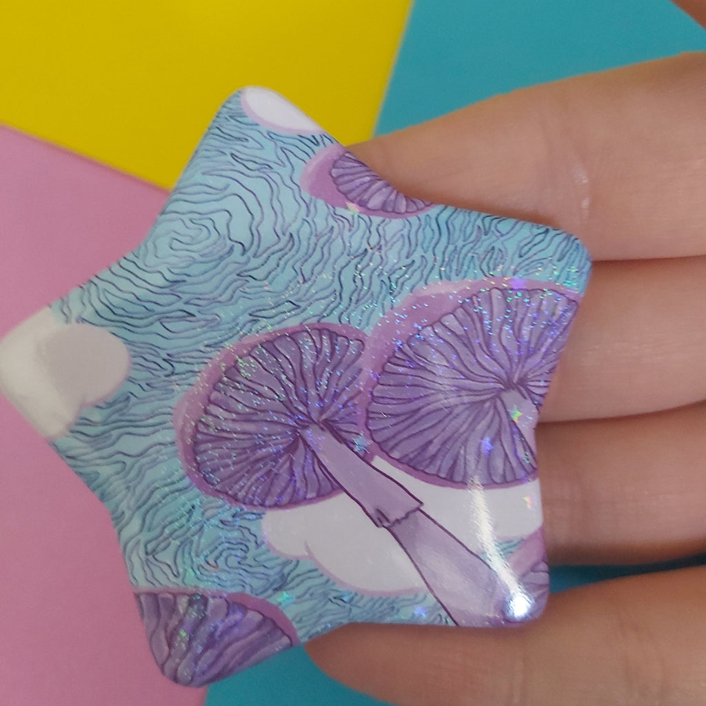 A hand holding a star-shaped badge featuring a purple and blue psychedelic mushroom pattern, with colourful paper backgrounds.
