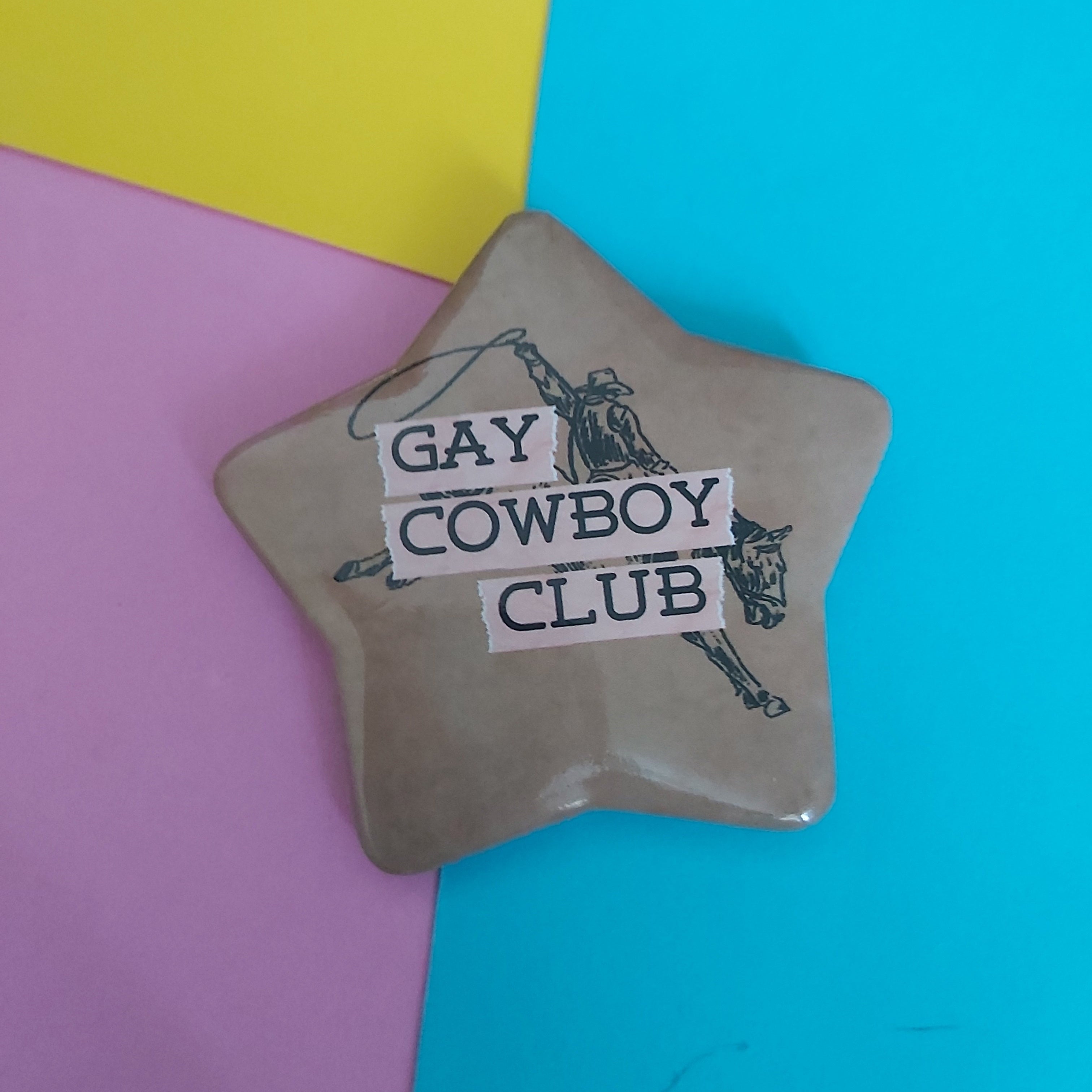 A star-shaped pin with a beige/tan background and black cowboy illustration, featuring the text 'GAY COWBOY CLUB' in white boxes. The pin is photographed on a background split diagonally into yellow, pink, and blue sections.