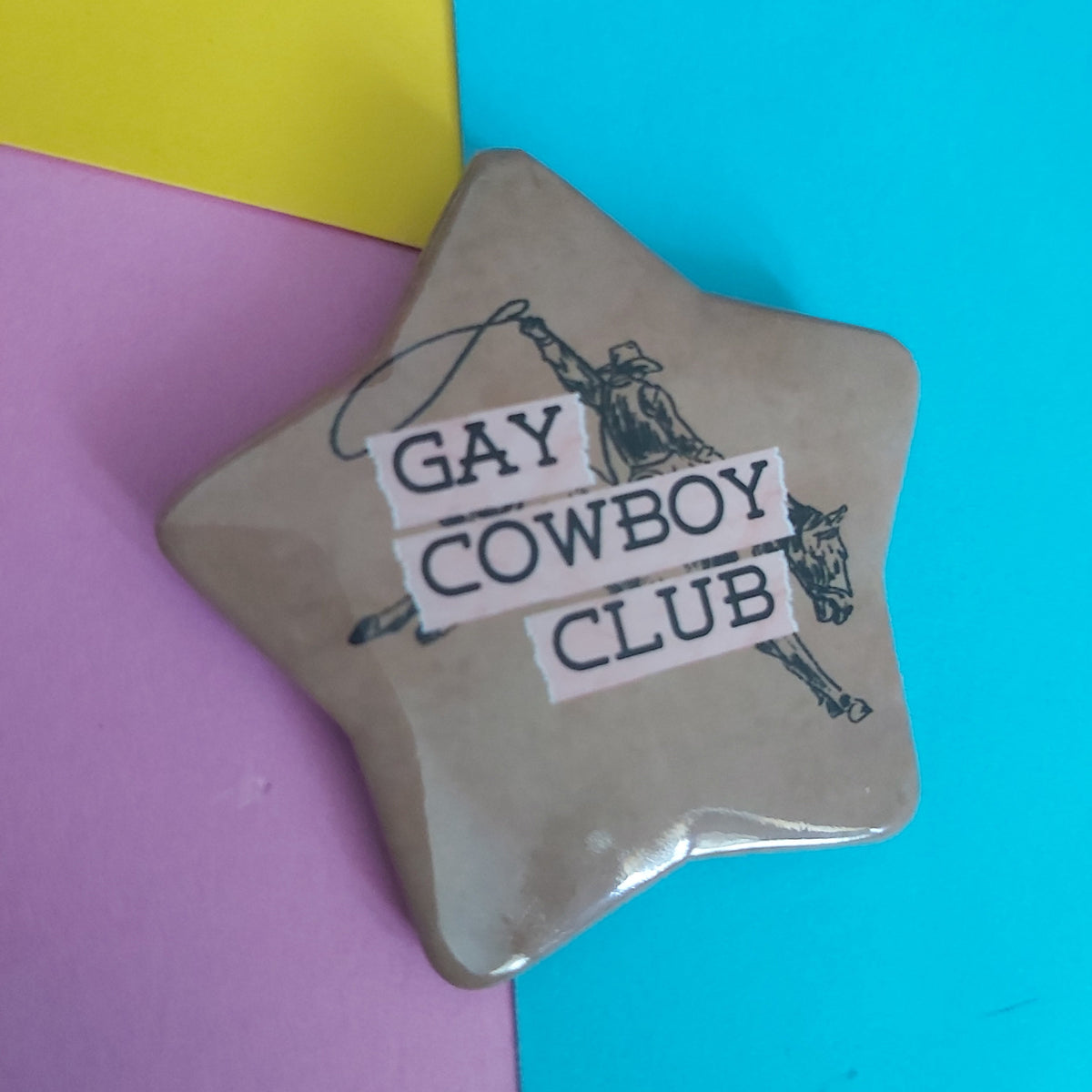 A star-shaped pin with a distressed tan background, featuring a black cowboy illustration and the text 'GAY COWBOY CLUB' in white lettering, set against a colourful background of yellow, pink, and blue paper.
