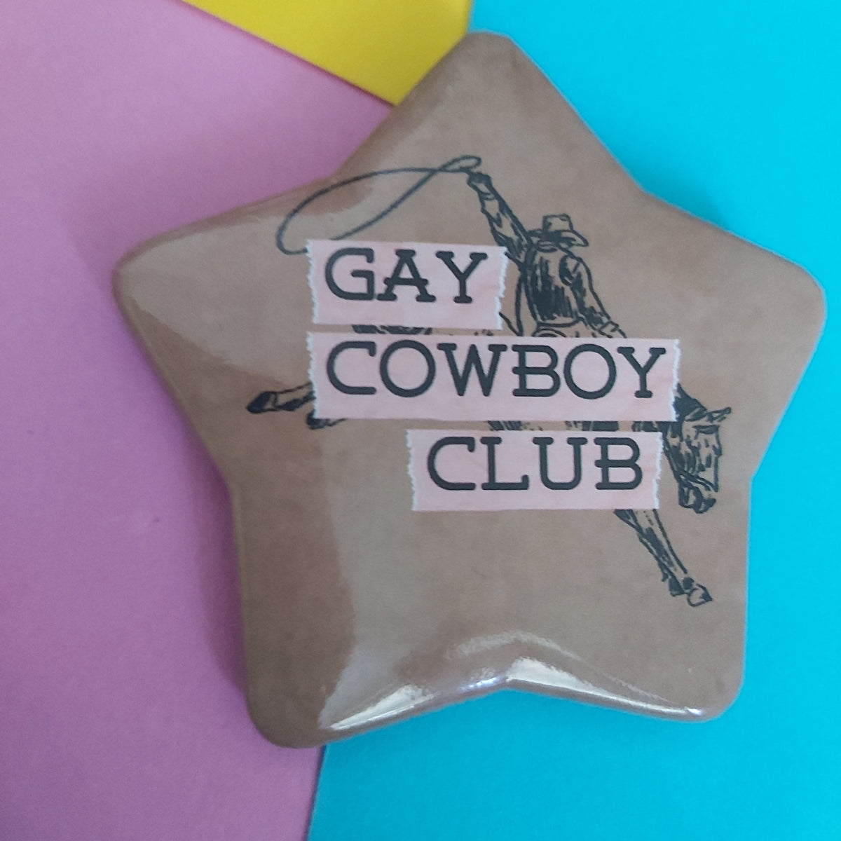 A star-shaped brown pin badge featuring a cowboy illustration and the text 'GAY COWBOY CLUB' in white lettering, placed on a colourful background of pink, yellow, and blue paper.