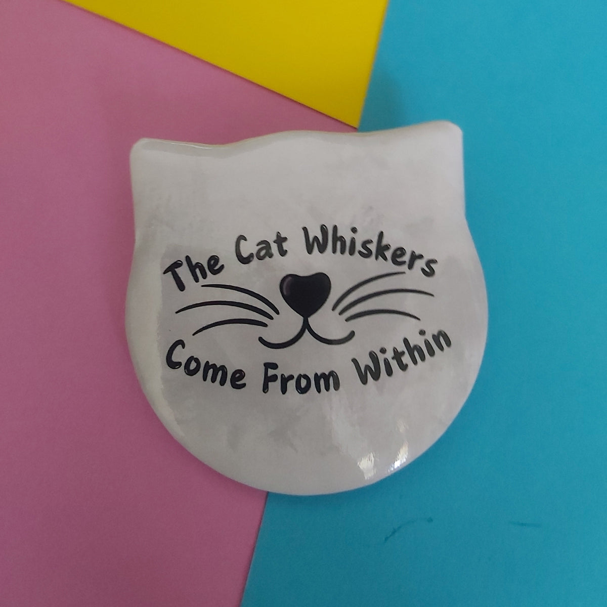 A white cat-shaped pin badge with black whisker lines and the phrase 'The Cat Whiskers Come From Within' printed on it, placed on a colourful background split diagonally into pink, yellow, and blue sections.