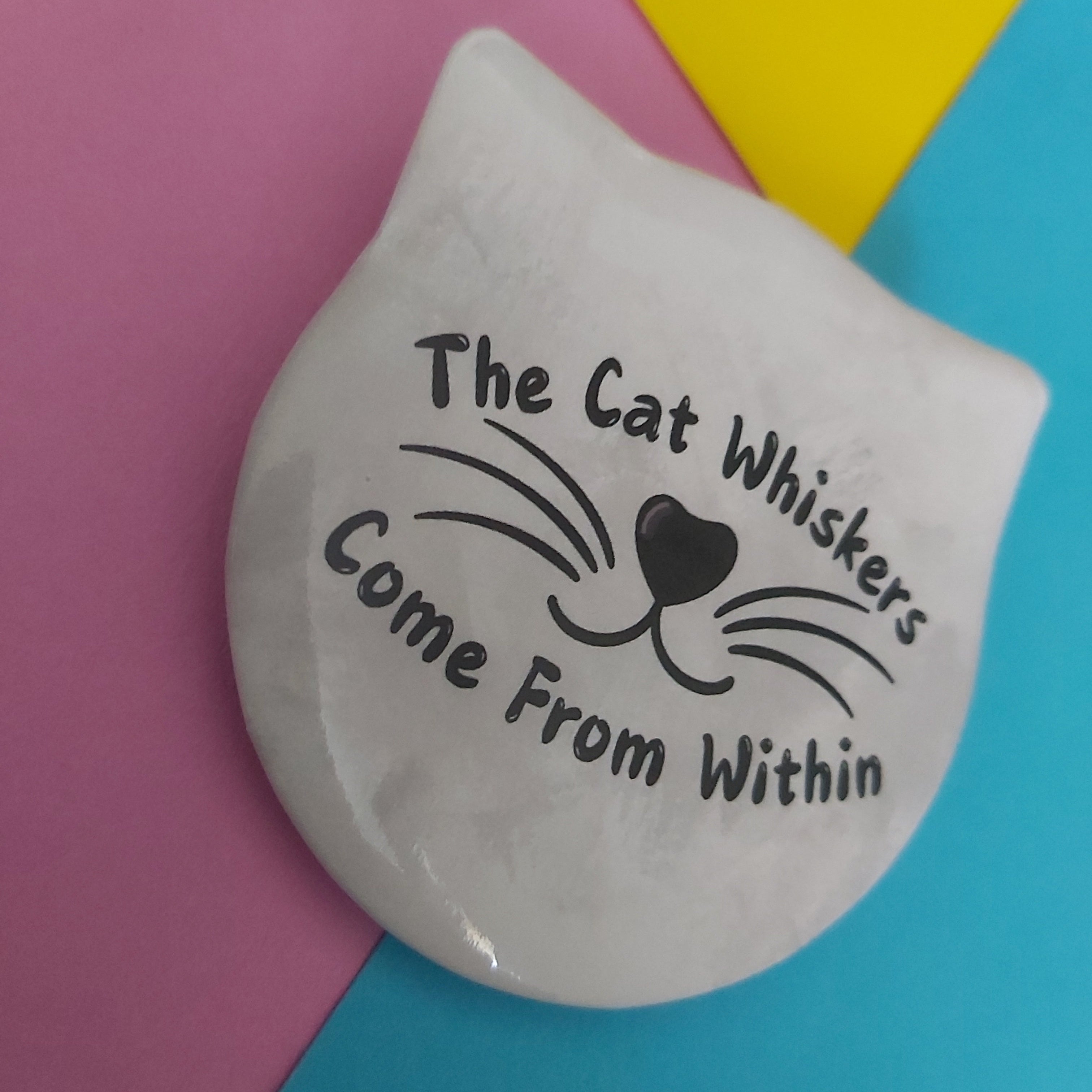 A white cat-shaped badge with black printed text reading 'The Cat Whiskers Come From Within' and a whisker-and-heart face design, placed on a pink, yellow, and blue colour-blocked background.