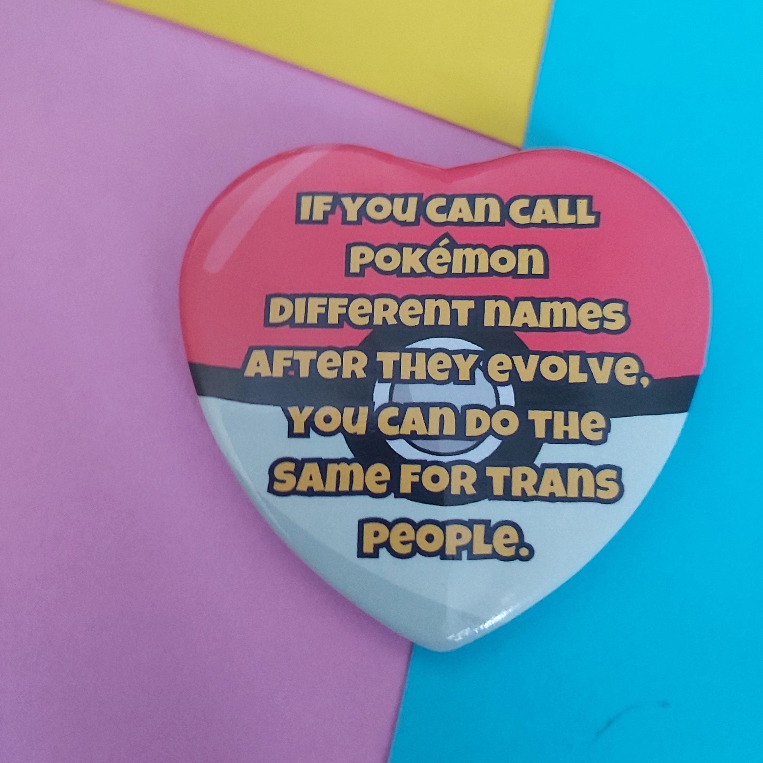 A heart-shaped pin badge styled like a Pokémon ball, featuring a red top half and a white bottom half with a black horizontal band, and the text reads, 'IF YOU CAN CALL POKÉMON DIFFERENT NAMES AFTER THEY EVOLVE, YOU CAN DO THE SAME FOR TRANS PEOPLE.