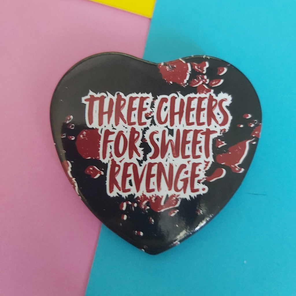 A heart-shaped black badge with red blood splatter details and the text 'THREE CHEERS FOR SWEET REVENGE' in white and red, displayed on a background split diagonally between pink, yellow, and blue.