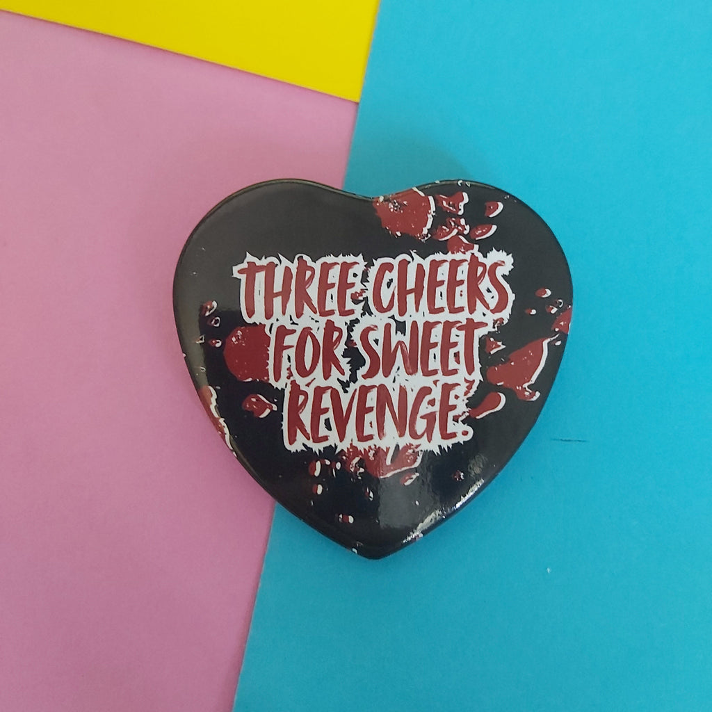 A heart-shaped black pin with red blood splatter details and the text 'THREE CHEERS FOR SWEET REVENGE' in white and red, displayed on a colourful background of pink, yellow, and blue paper.