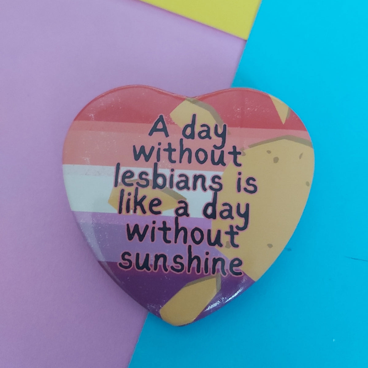 A heart-shaped pinback button with a striped design and the text 'A day without lesbians is like a day without sunshine', displayed on a colourful background of purple, yellow, and blue paper.
