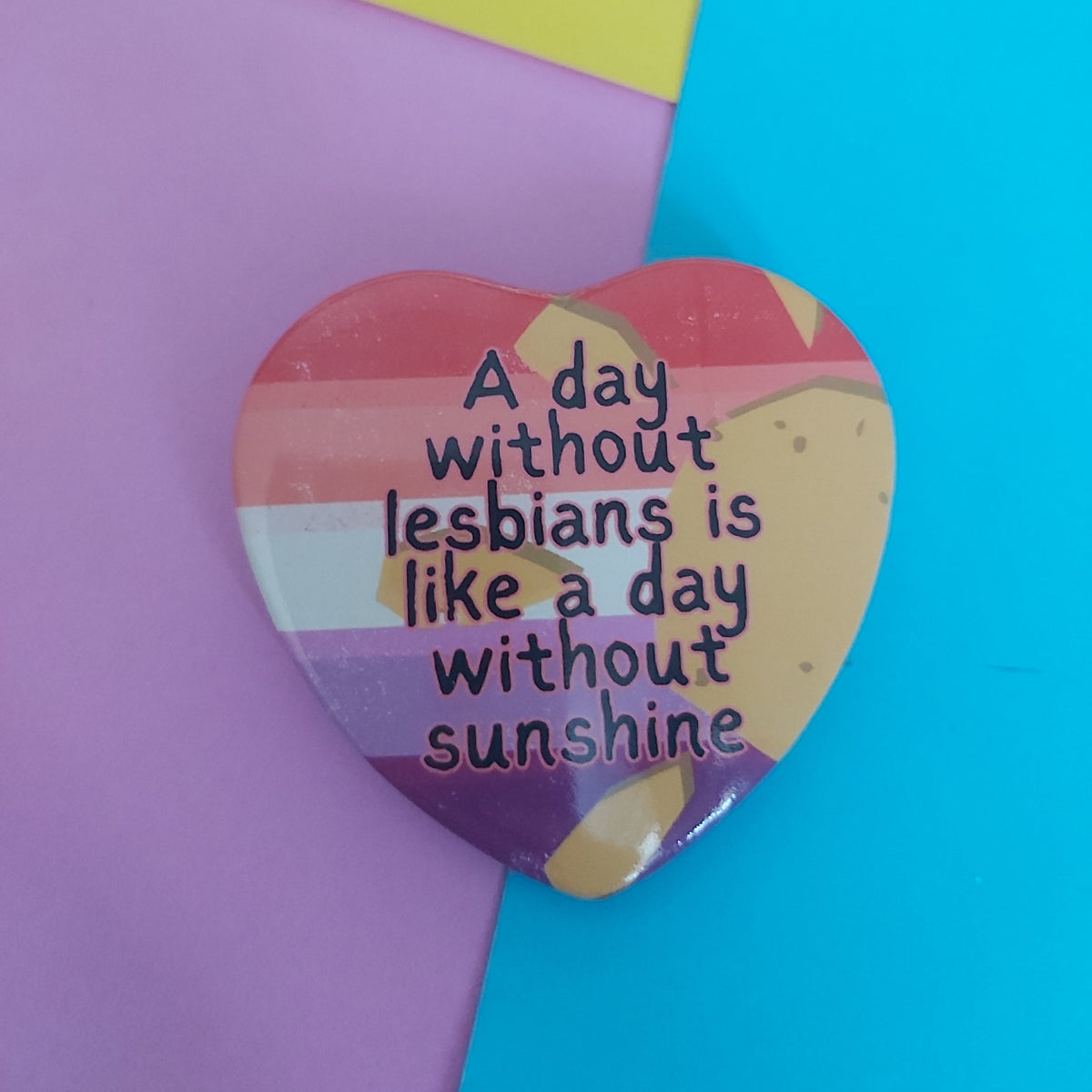 A heart-shaped pin with a colourful striped and sun motif, featuring the text 'A day without lesbians is like a day without sunshine' in black lettering. The pin is displayed on a background split diagonally between pastel purple and bright cyan blue.
