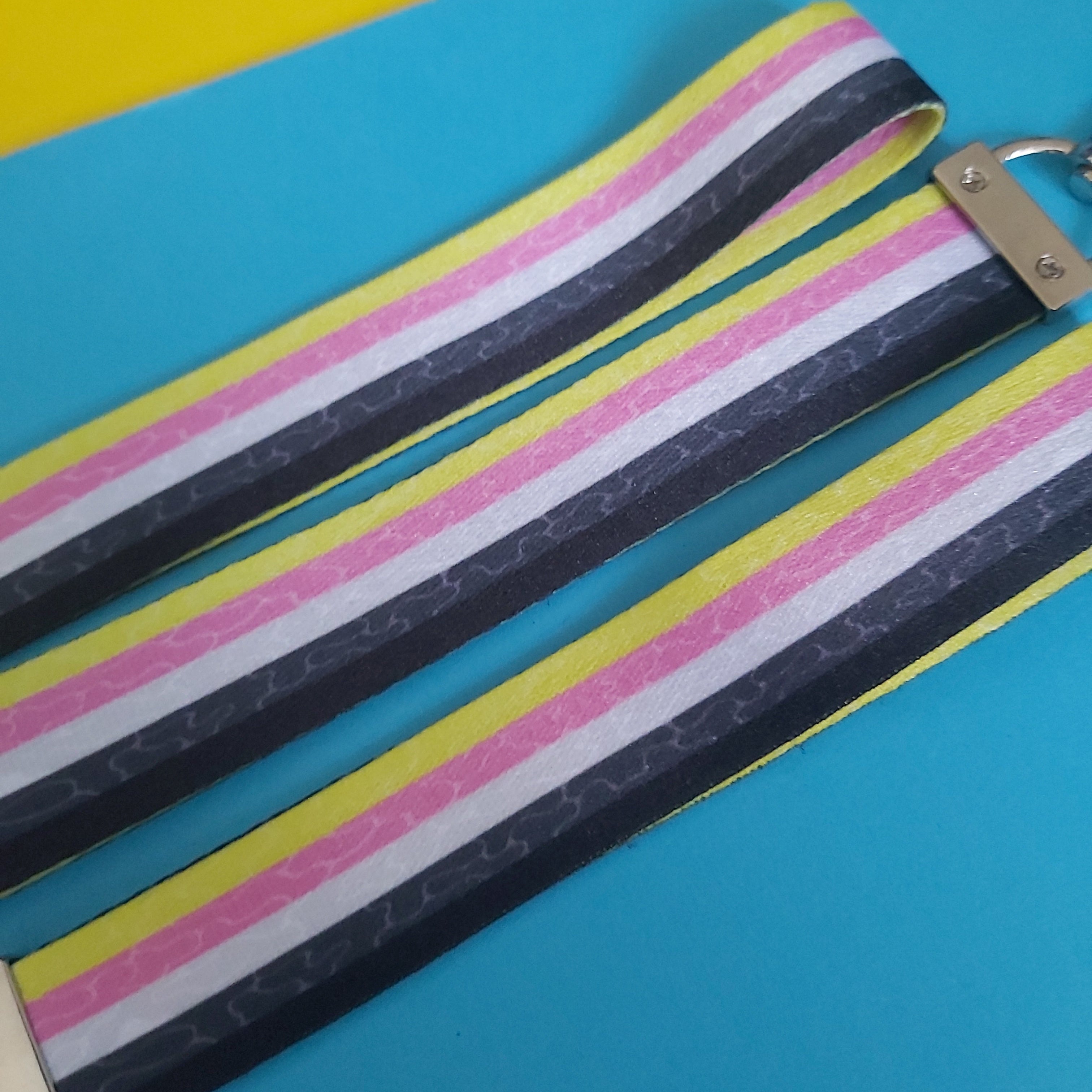 A close-up of a striped fabric lanyard with a metal lobster clasp, laid diagonally on a bright blue background with a yellow corner visible.