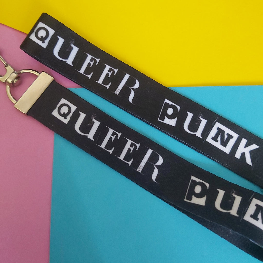 A black fabric lanyard strap with the words 'QUEER PUNK' printed in white stencil-style lettering, attached to a gold-tone metal clasp, laid diagonally across a colorful background of yellow, pink, and blue paper.