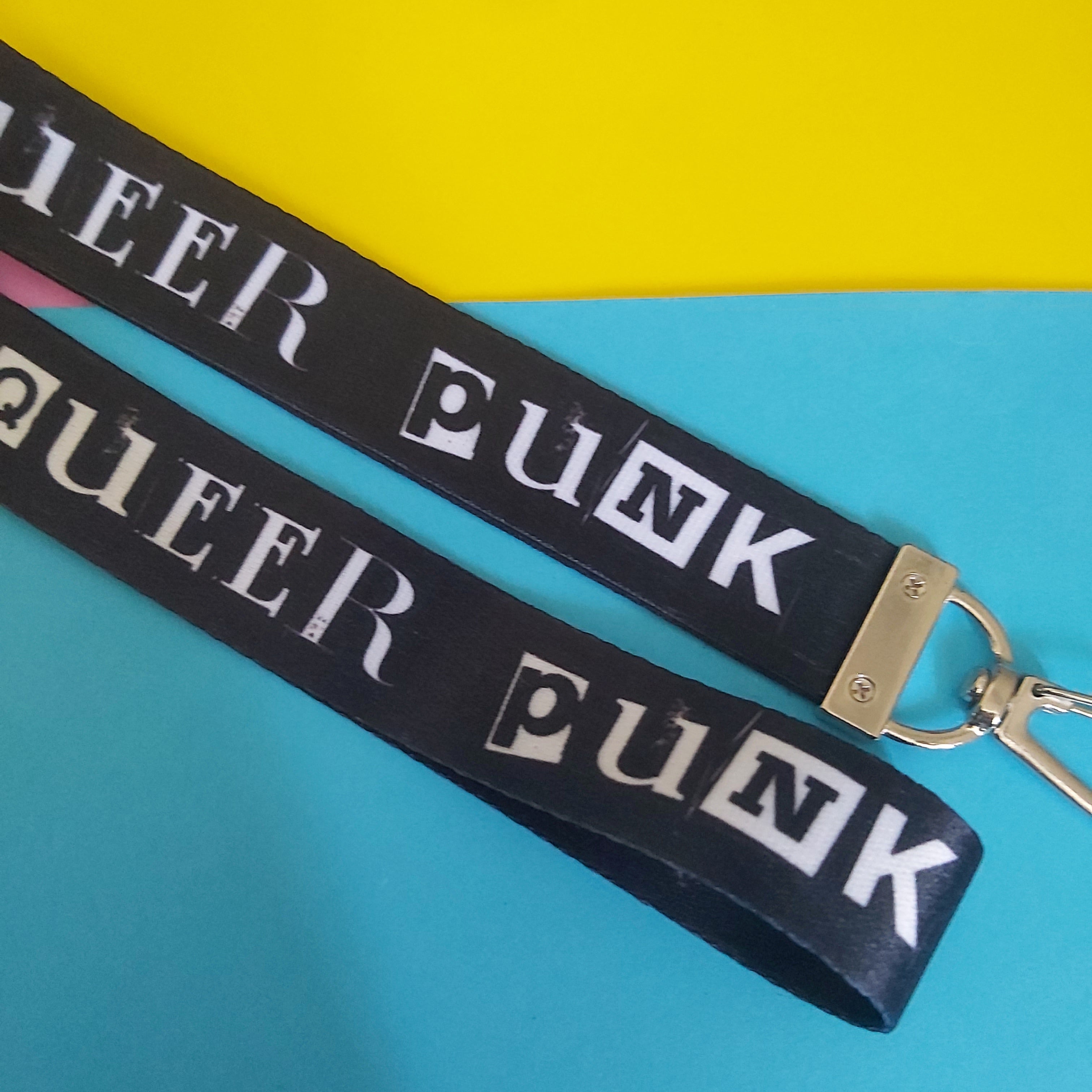 A black fabric wrist lanyard with the words 'QUEER PUNK' printed repeatedly in white block letters, featuring a silver metal clasp, laid diagonally on a split yellow and blue background.