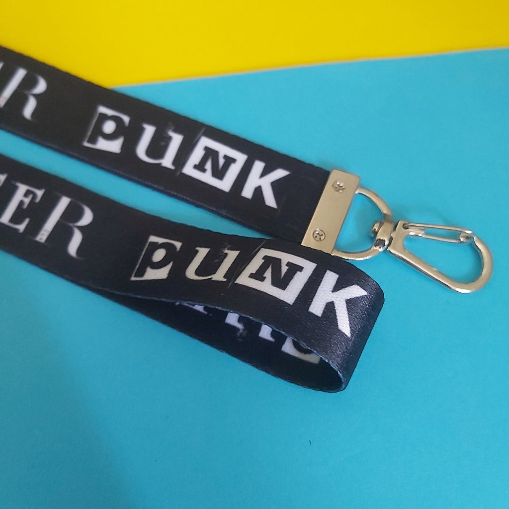A black woven lanyard strap with repeating white 'PUNK' lettering and a silver metal lobster clasp, photographed on a bright blue and yellow background.