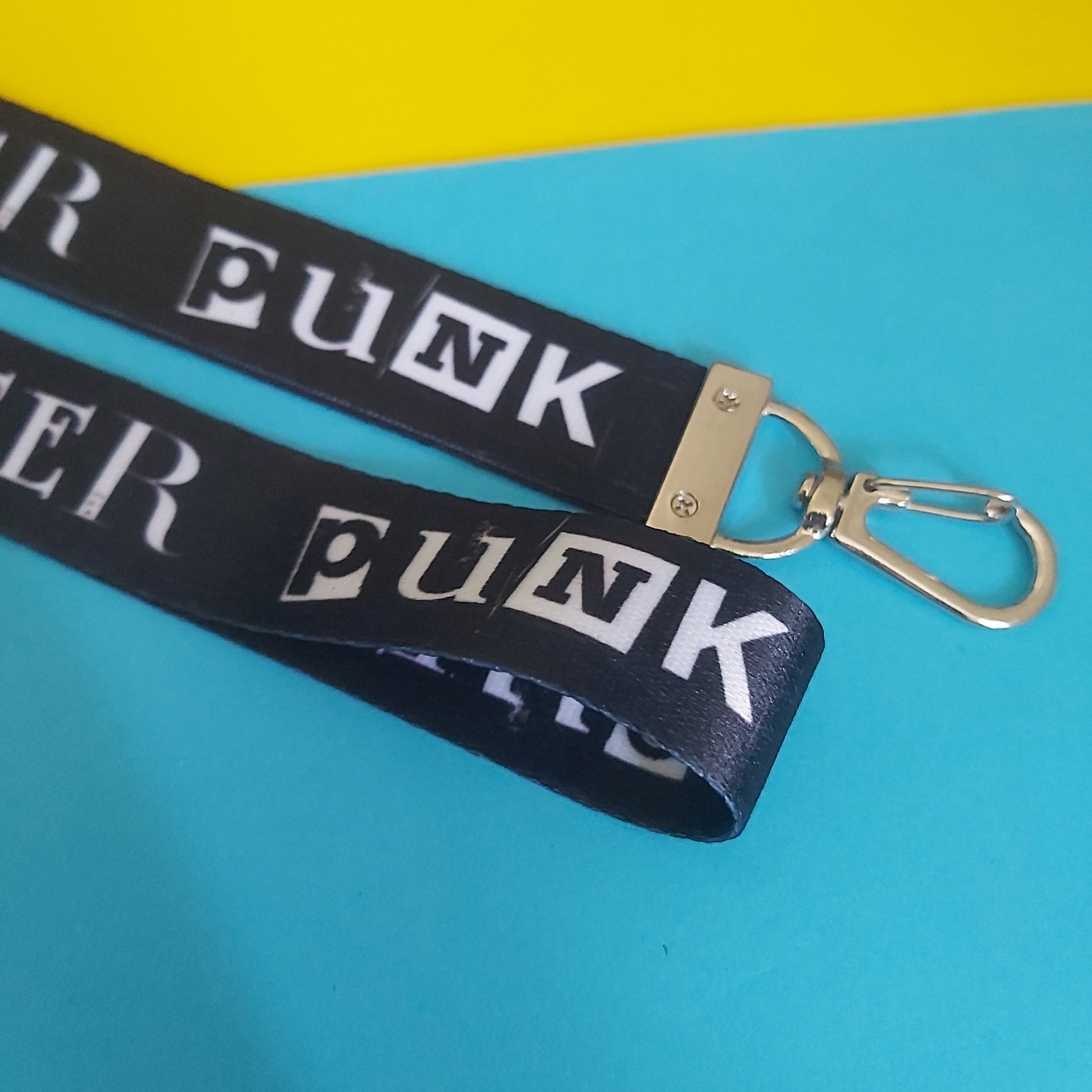 A black woven lanyard strap with repeating white 'PUNK' lettering and a silver metal lobster clasp, photographed on a bright blue and yellow background.