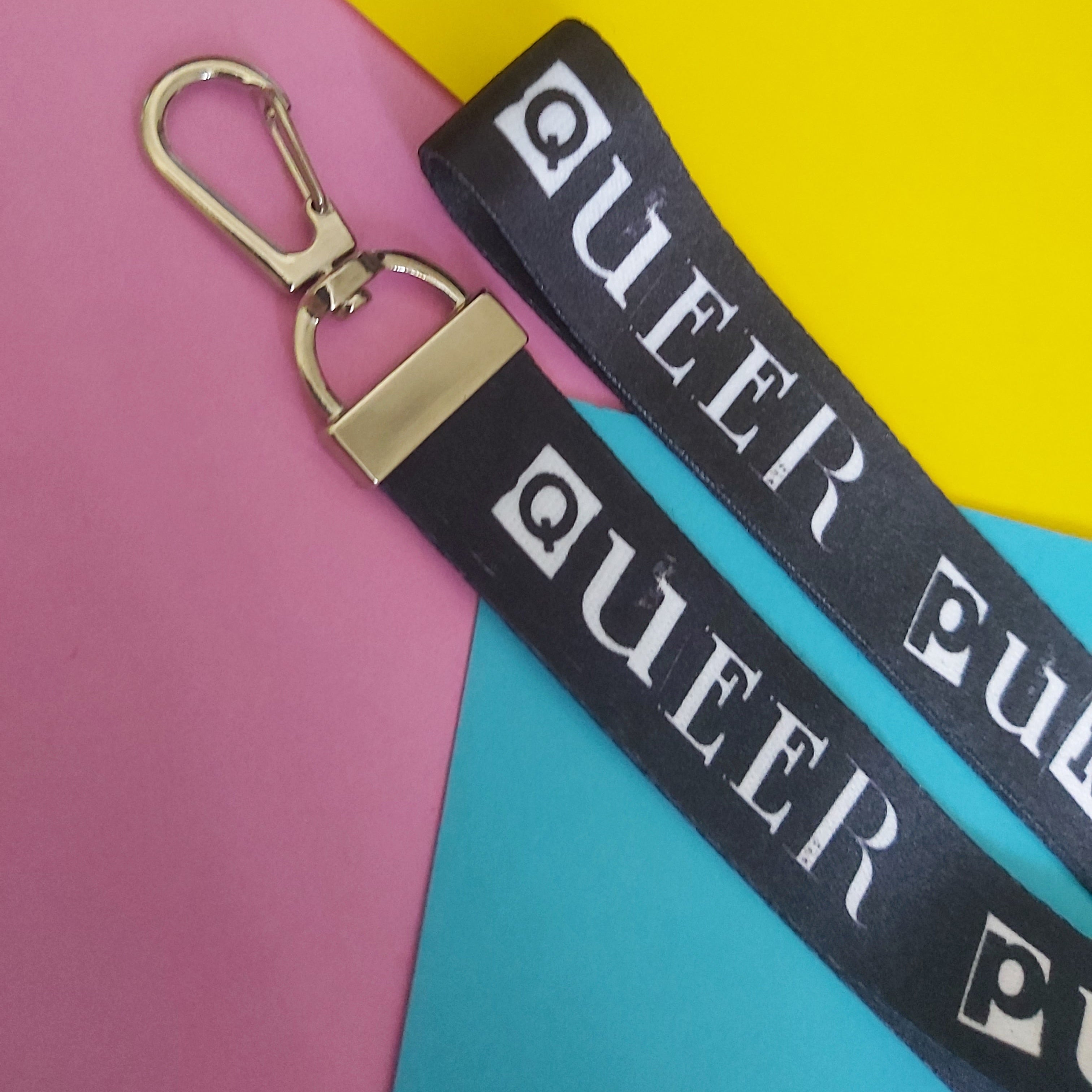 A black fabric keychain lanyard strap with a silver carabiner clip, printed with the word 'QUEER PUNK' in bold white letters, laid diagonally across a colourful background of pink, yellow, and blue paper.