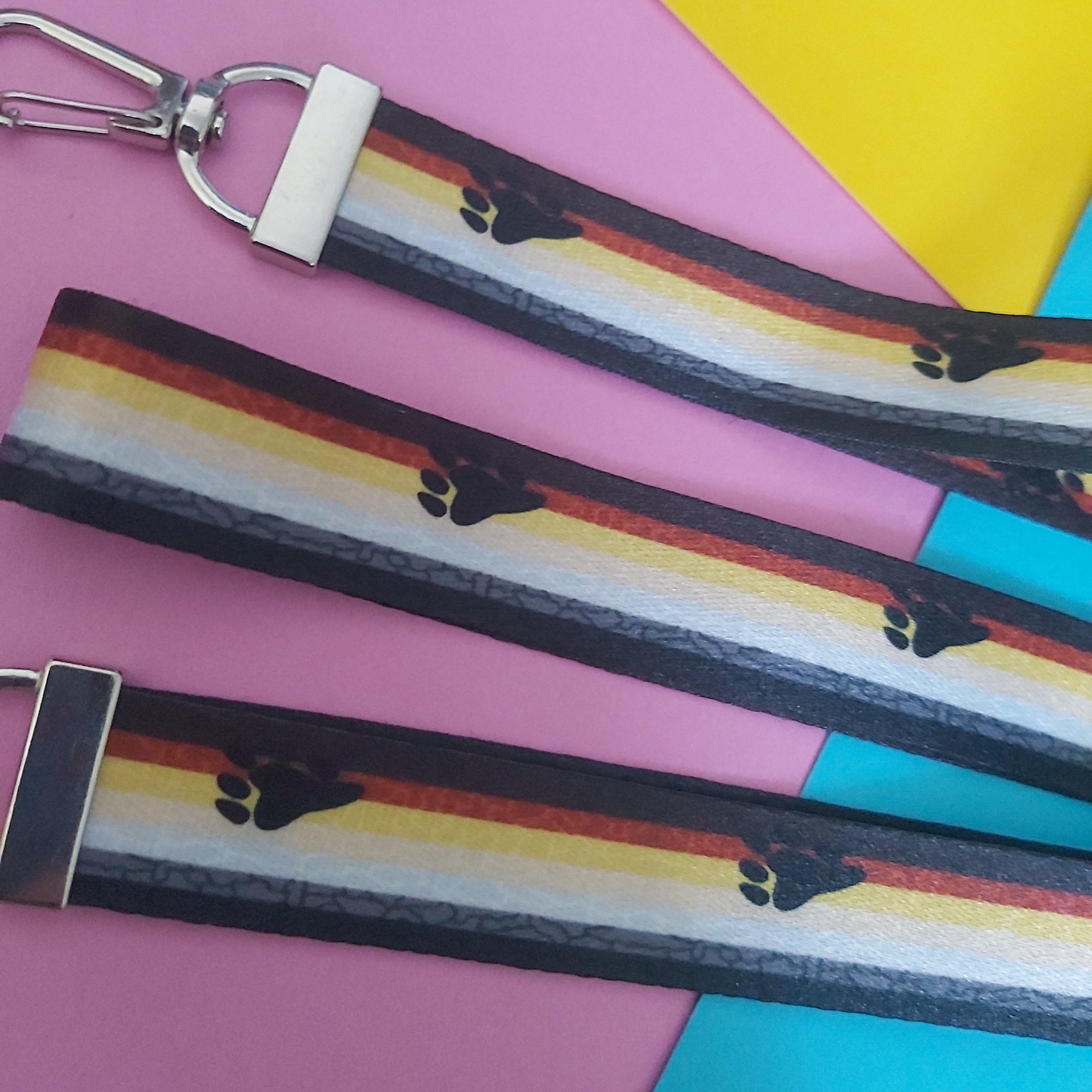 A close-up of three matching printed straps laid diagonally on a colorful pink, yellow, and blue background. The straps have a black, orange, yellow, white, and grey striped design with small black paw prints, plus silver metal clips and white plastic connectors.