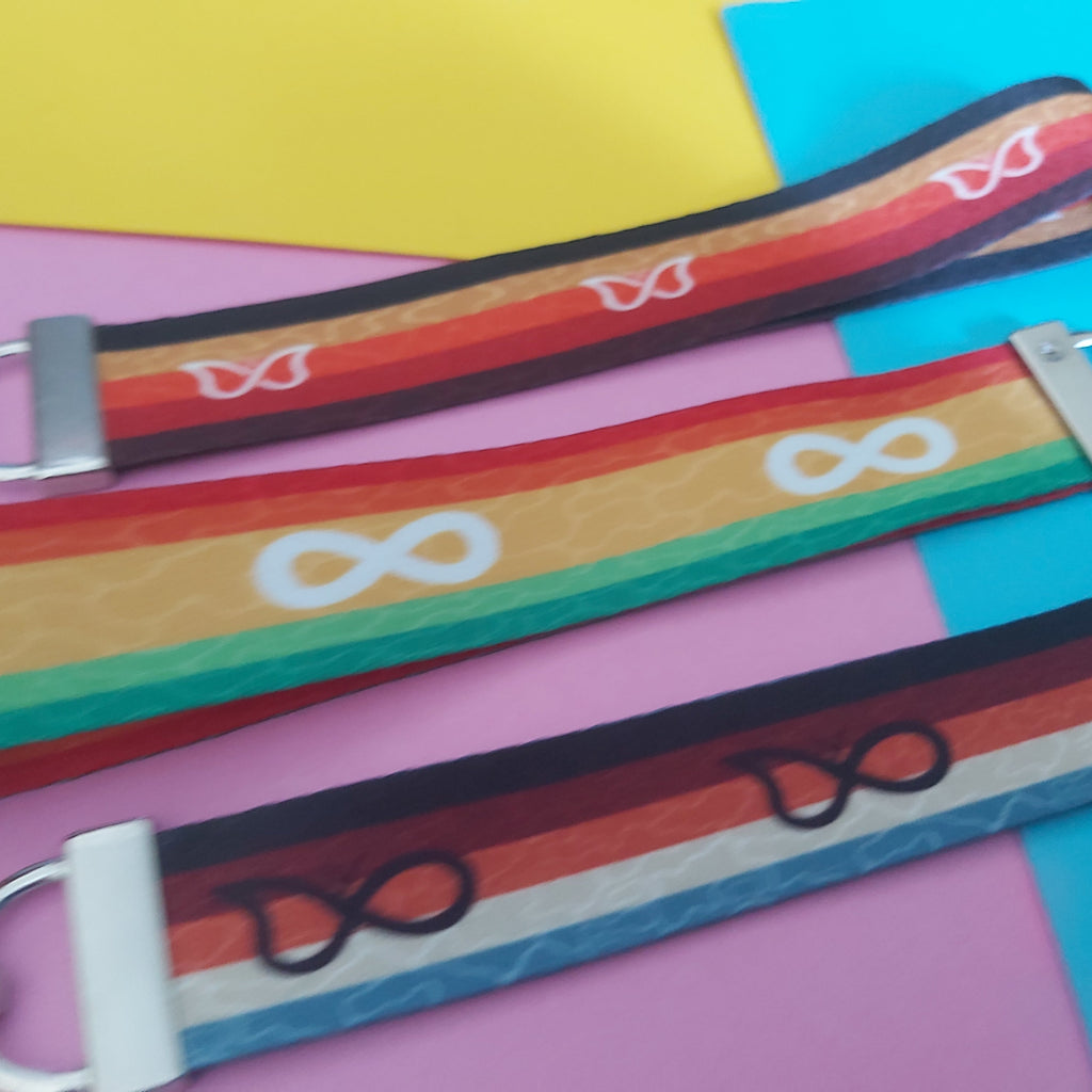 Three colorful fabric keychains with silver metal clips, laid diagonally on a background of pink, yellow, and blue paper. The keychains feature rainbow stripes and white symbols including infinity signs.
