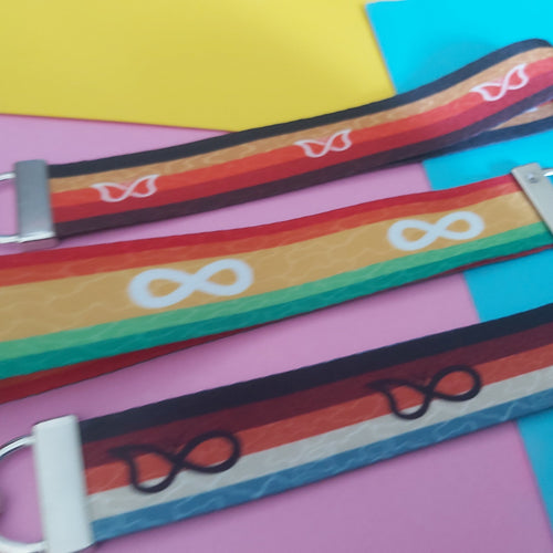 Three colorful fabric keychains with silver metal clips, laid diagonally on a background of pink, yellow, and blue paper. The keychains feature rainbow stripes and white symbols including infinity signs.