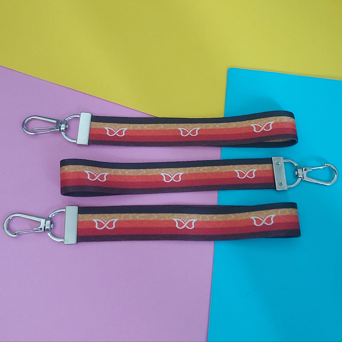 Three fabric keychains with silver swivel clips, laid flat on a colourful background of yellow, blue, and pink paper. The keychains feature a striped pattern in red, orange, and brown with white butterfly motifs.