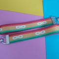 A close-up of a fabric keychain bag strap with a yellow centre, red and green stripes on the sides, and white infinity symbols printed repeatedly. The strap is laid diagonally across sheets of colored paper in yellow, pink, and blue.