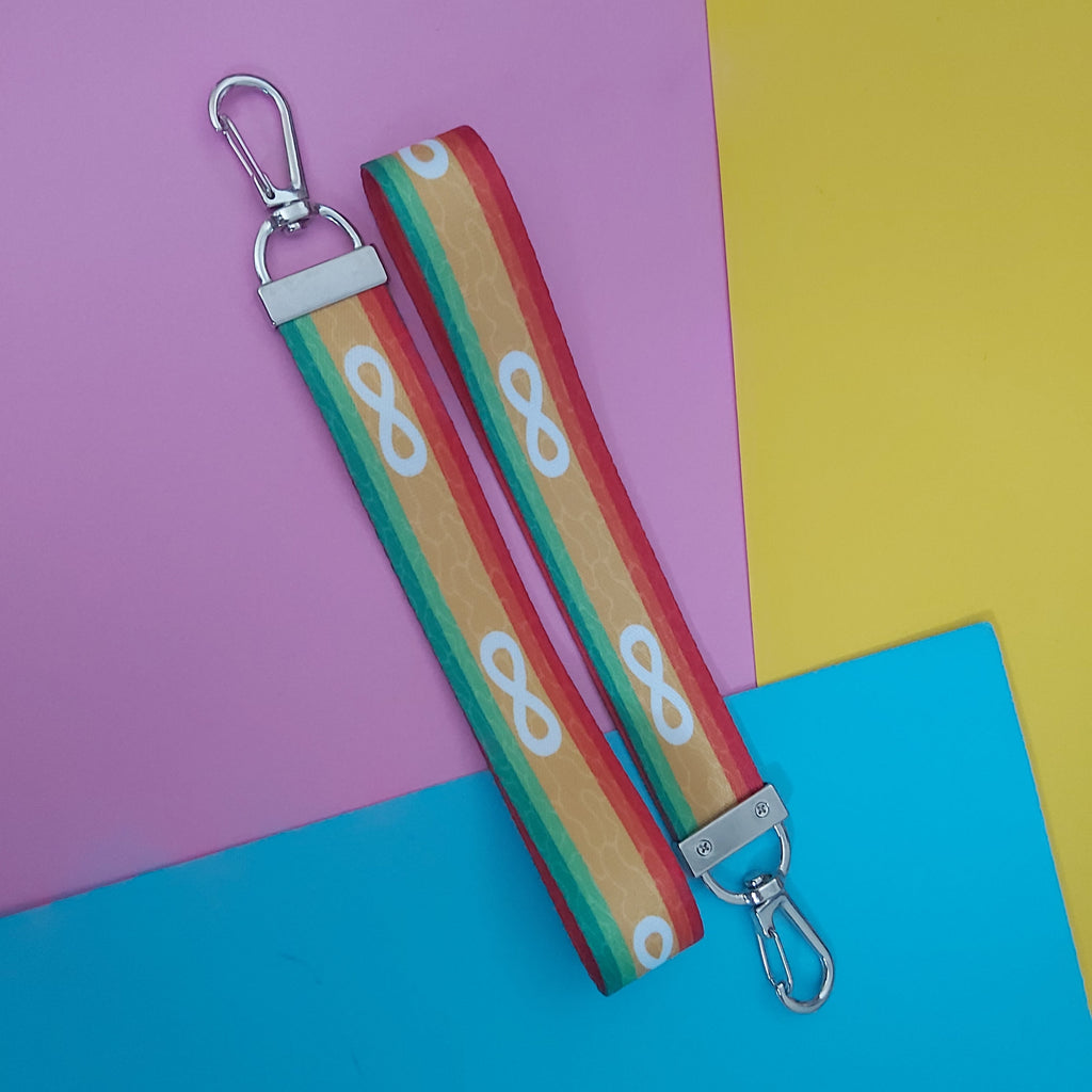 A fabric keychain bag strap with a colourful striped pattern in red, orange, yellow, green, featuring white infinity symbols. It has silver metal swivel clips on both ends and is displayed diagonally on a split pink, yellow, and blue background.