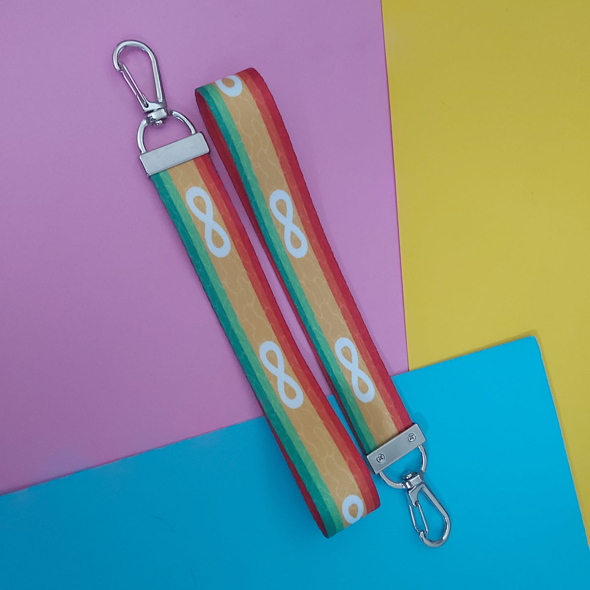 A fabric keychain bag strap with a colourful striped pattern in red, orange, yellow, green, featuring white infinity symbols. It has silver metal swivel clips on both ends and is displayed diagonally on a split pink, yellow, and blue background.