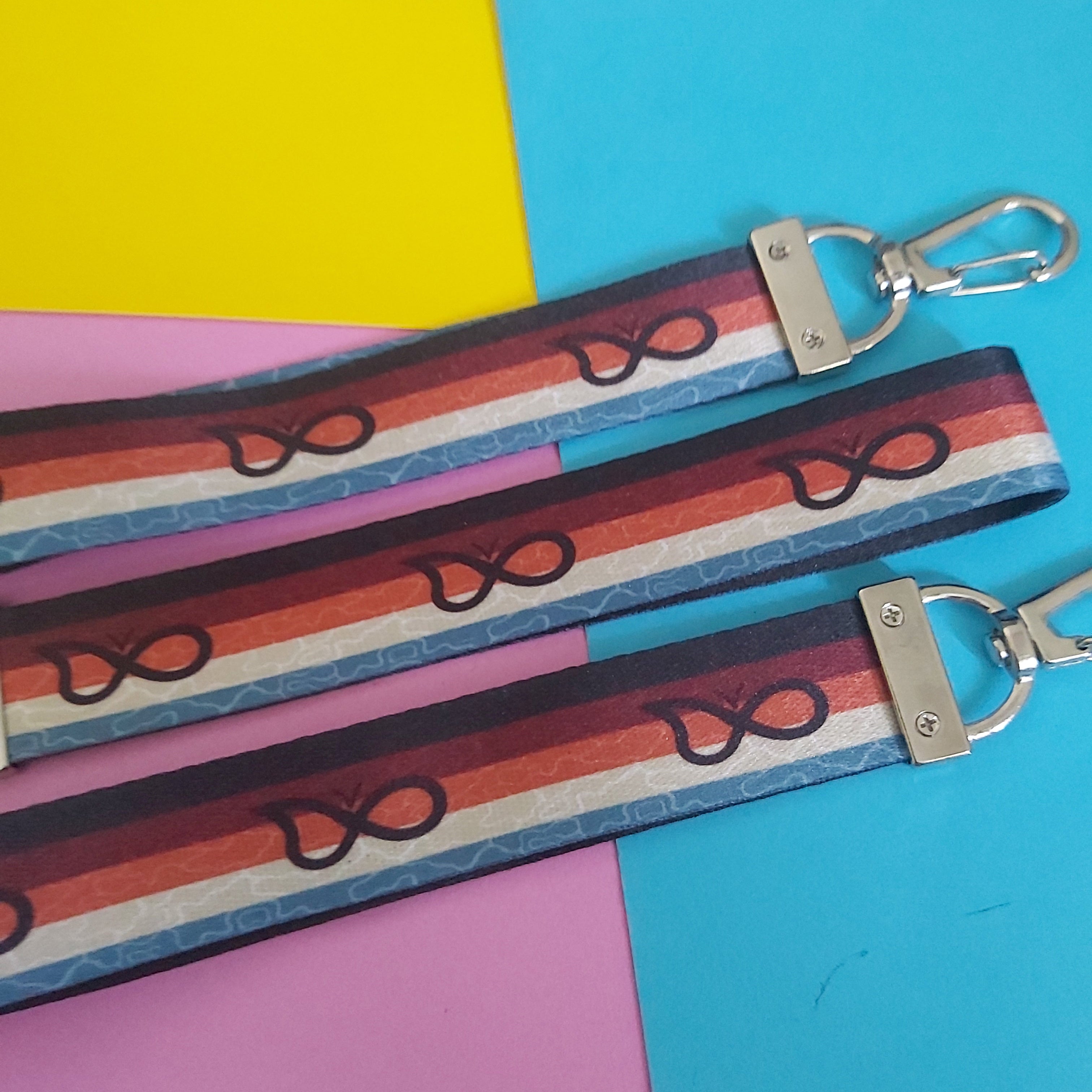 Three fabric keychains with silver metal clasp hardware, featuring a retro striped pattern in red, orange, blue, and white laid flat on a colourful yellow, blue, and pink background.